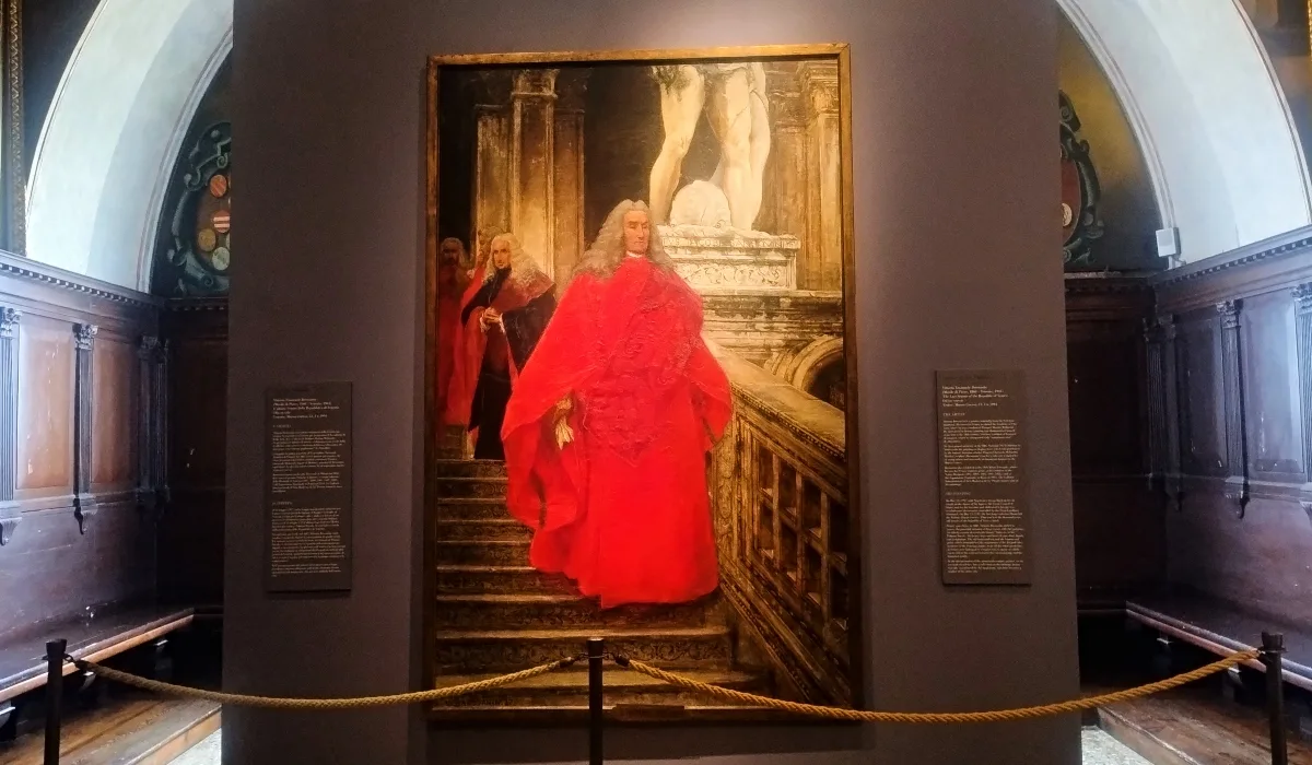 Pintura titulada “The Dux” de John Everett Millais, mostrando a un dux veneciano con túnica roja descendiendo por una escalera de mármol en el Palacio Ducal de Venecia, con otros dignatarios al fondo y esculturas clásicas detrás.