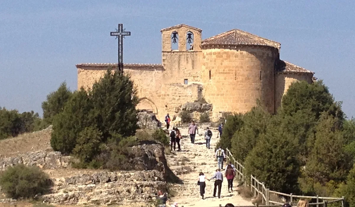 Ermita románica de San Frutos en lo alto de un risco, con visitantes subiendo por un camino de piedra entre árboles, en el Parque Natural de las Hoces del Río Duratón, Segovia.