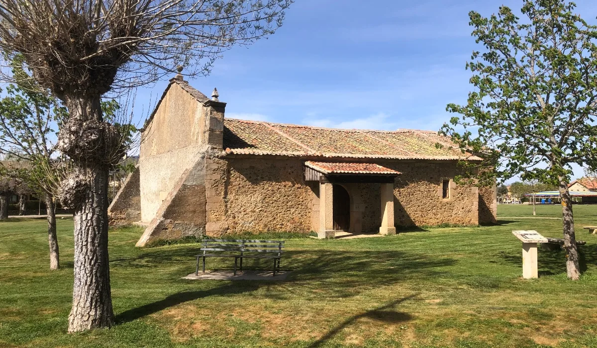 Ermita de San Roque en Riaza, Segovia, edificio de piedra con tejado de tejas situado en un prado verde con árboles y bancos bajo un cielo azul despejado