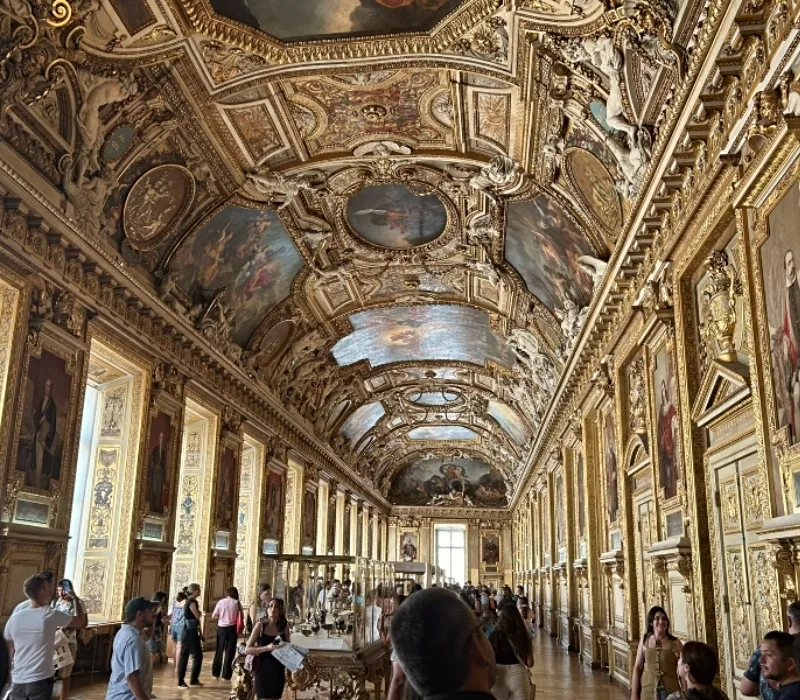 Vista interior de la Galería de Apolo en el Museo del Louvre. El salón es alargado, con techos altos abovedados ricamente decorados con molduras doradas, frescos y esculturas. Las paredes están adornadas con retratos enmarcados y ornamentación dorada. Varios visitantes recorren la sala, observando vitrinas con objetos expuestos en el centro.