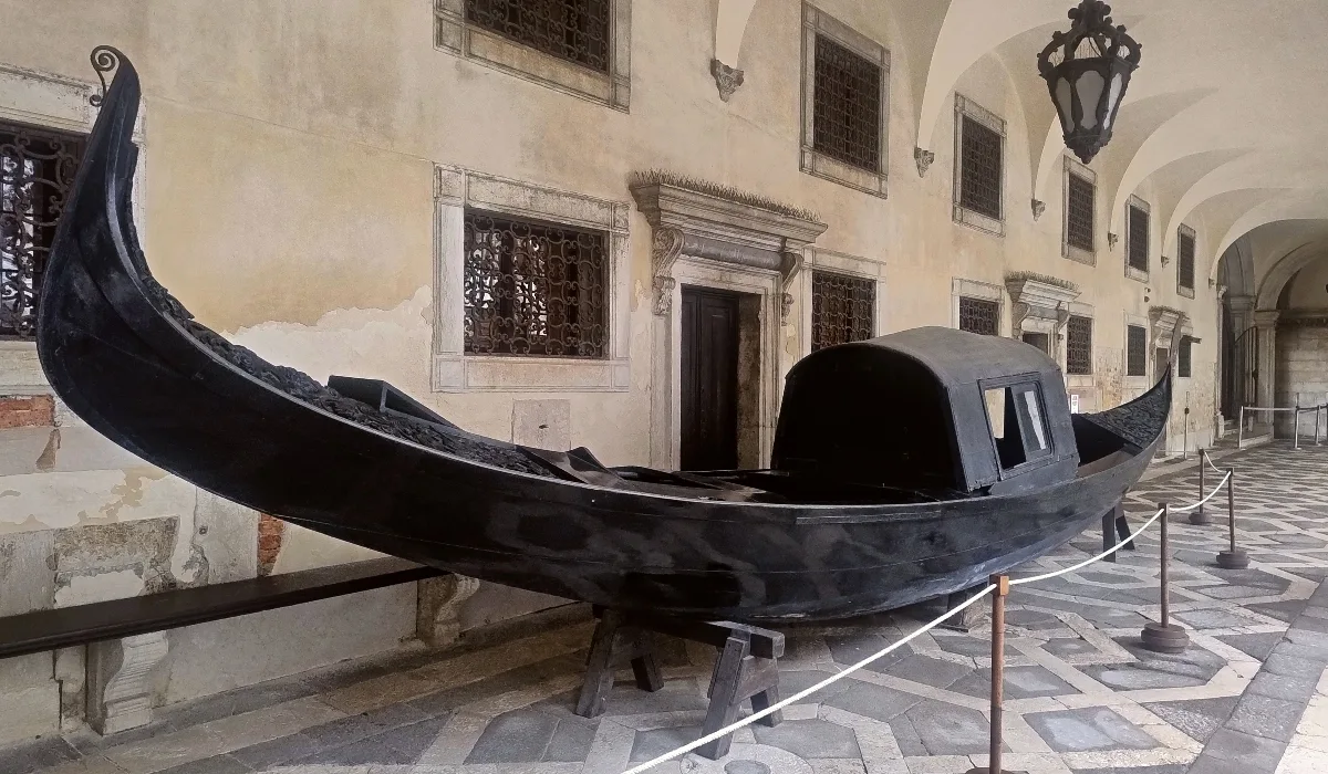 Antigua góndola veneciana negra expuesta bajo las arcadas del Palacio Ducal, junto a ventanas enrejadas y lámparas de hierro forjado.
