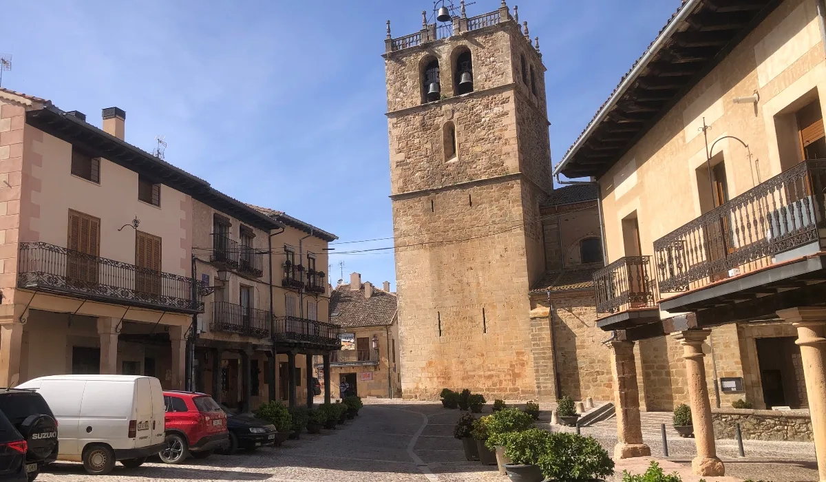 Ermita de Nuestra Señora del Manto en Riaza, Segovia, con torre campanario de piedra y entorno de casas tradicionales con balcones de madera y calles empedradas