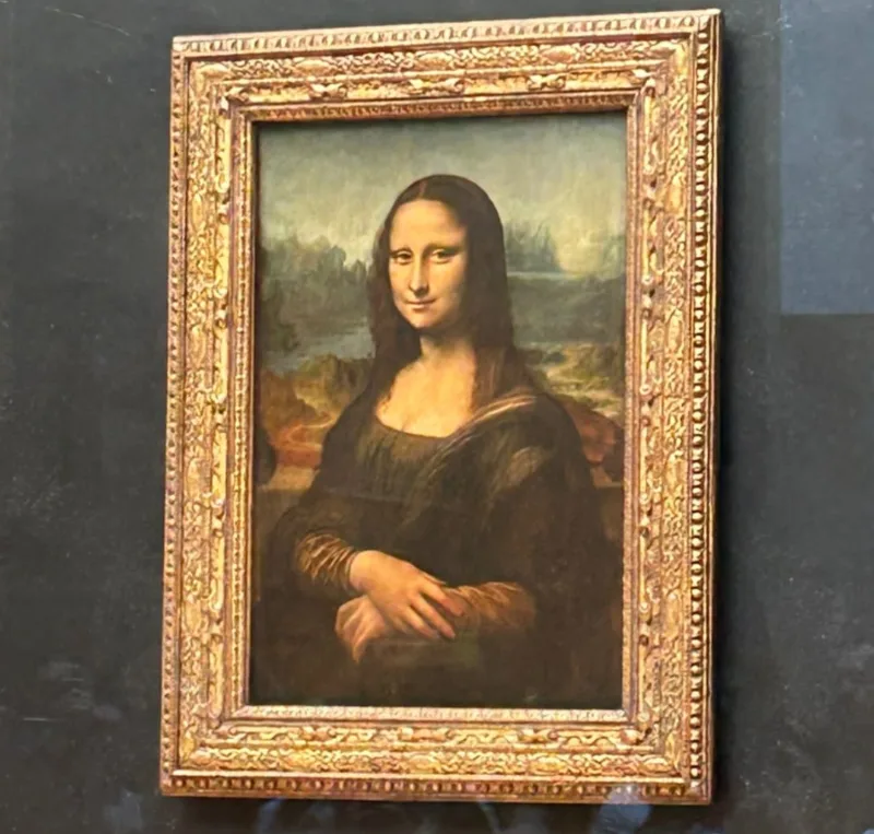 Pintura de la Mona Lisa, retrato renacentista de una mujer con expresión enigmática, cabello oscuro y vestido sobrio, enmarcada en un marco dorado ornamentado y expuesta tras un cristal.