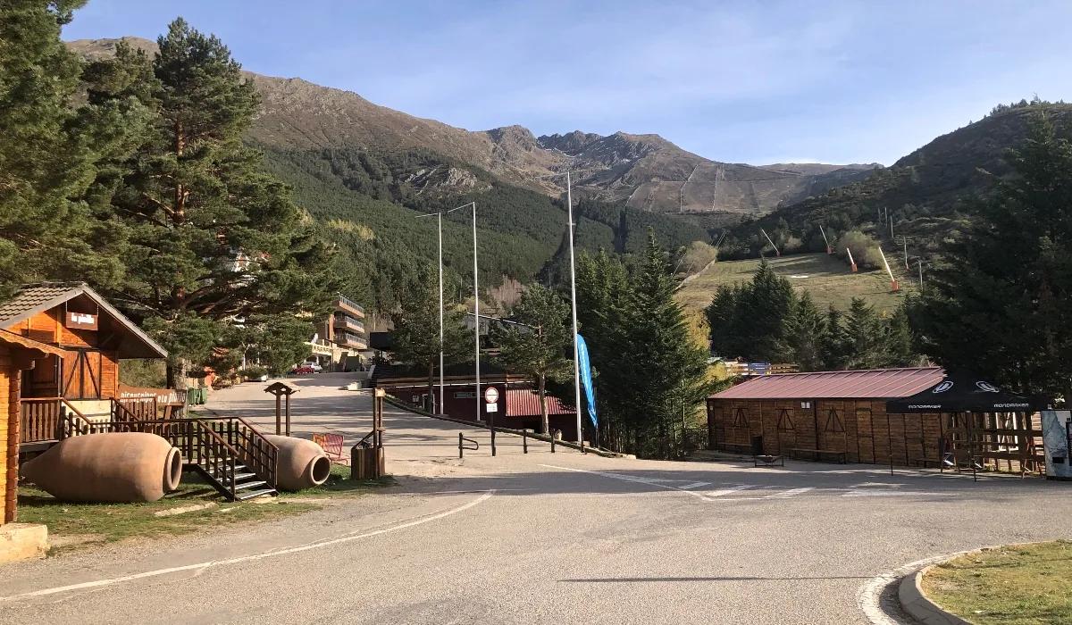 Vista de la estación de esquí de La Pinilla, en la provincia de Segovia, con montañas al fondo, pistas de esquí visibles, árboles y edificaciones de madera en primer plano bajo un cielo despejado.