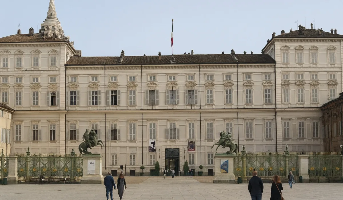 Fachada del Palazzo Reale di Torino en la Piazza Castello, con la bandera italiana ondeando sobre el edificio y algunas personas caminando por la plaza en un día despejado