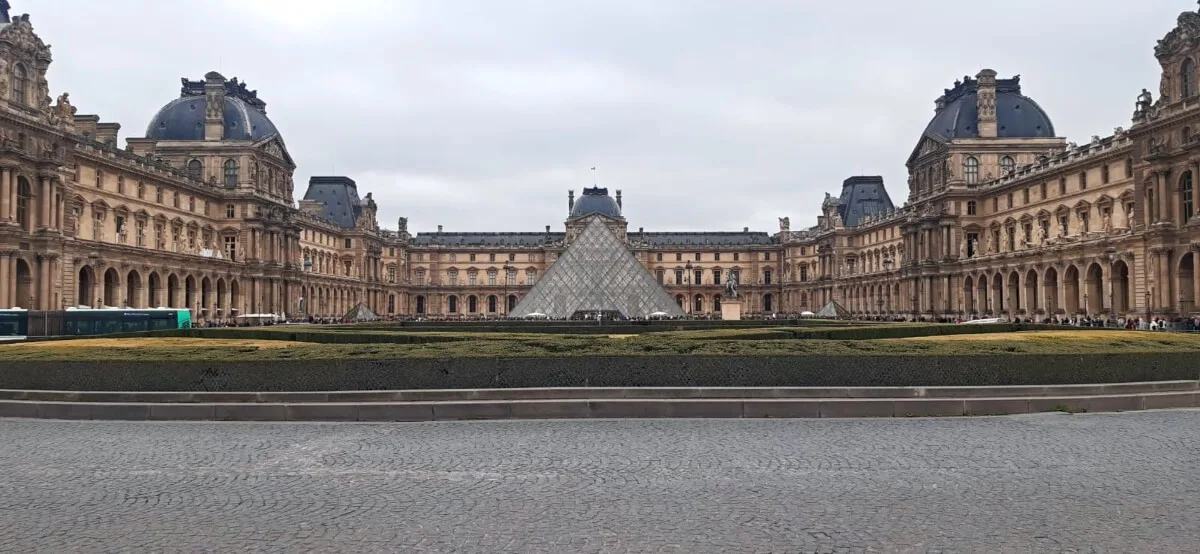 Vista frontal del Museo del Louvre en París, con la icónica pirámide de cristal en el centro del patio rodeada por el histórico palacio de estilo renacentista bajo un cielo nublado