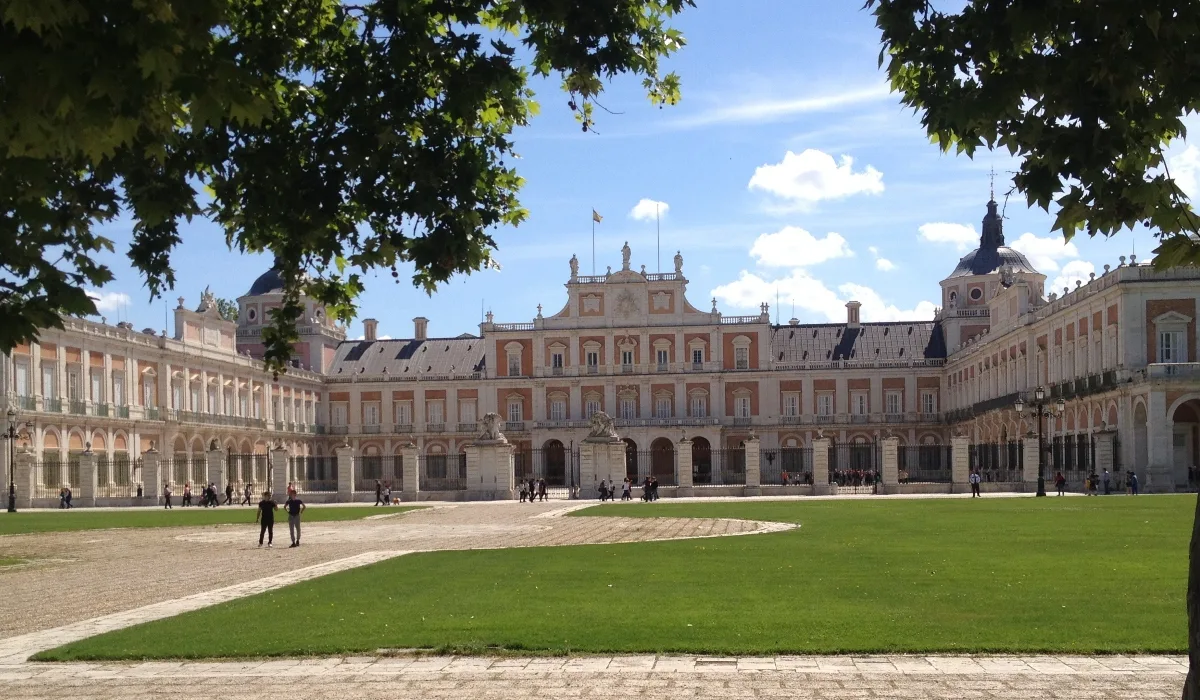 Fachada principal del Palacio Real de Aranjuez, con amplios jardines y visitantes paseando bajo un cielo despejado