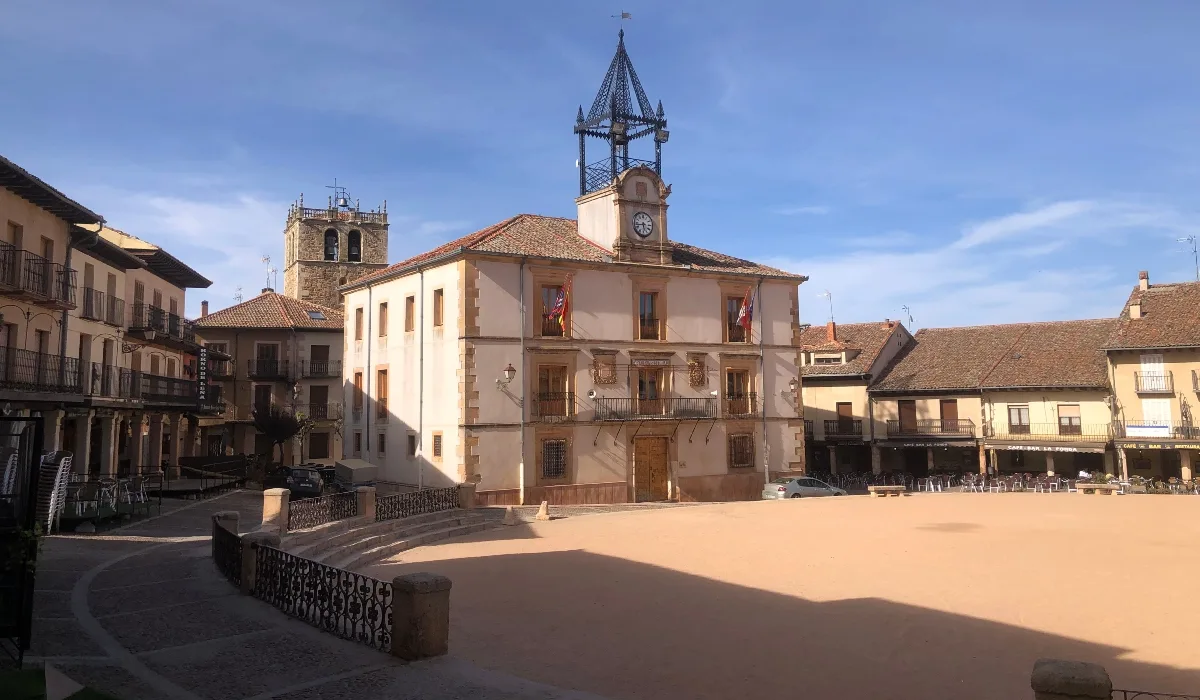 Vista de la Plaza Mayor de Riaza, Segovia, con el edificio del Ayuntamiento en el centro, una torre con reloj y banderas, rodeado de casas con balcones típicos castellanos y un cielo despejado