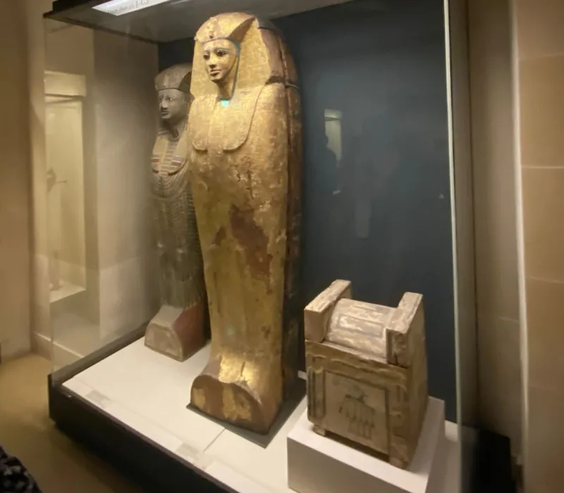 Sarcófago antropomorfo egipcio en exposición en el Museo del Louvre, decorado con detalles dorados y acompañado de otro sarcófago y un cofre canópico.