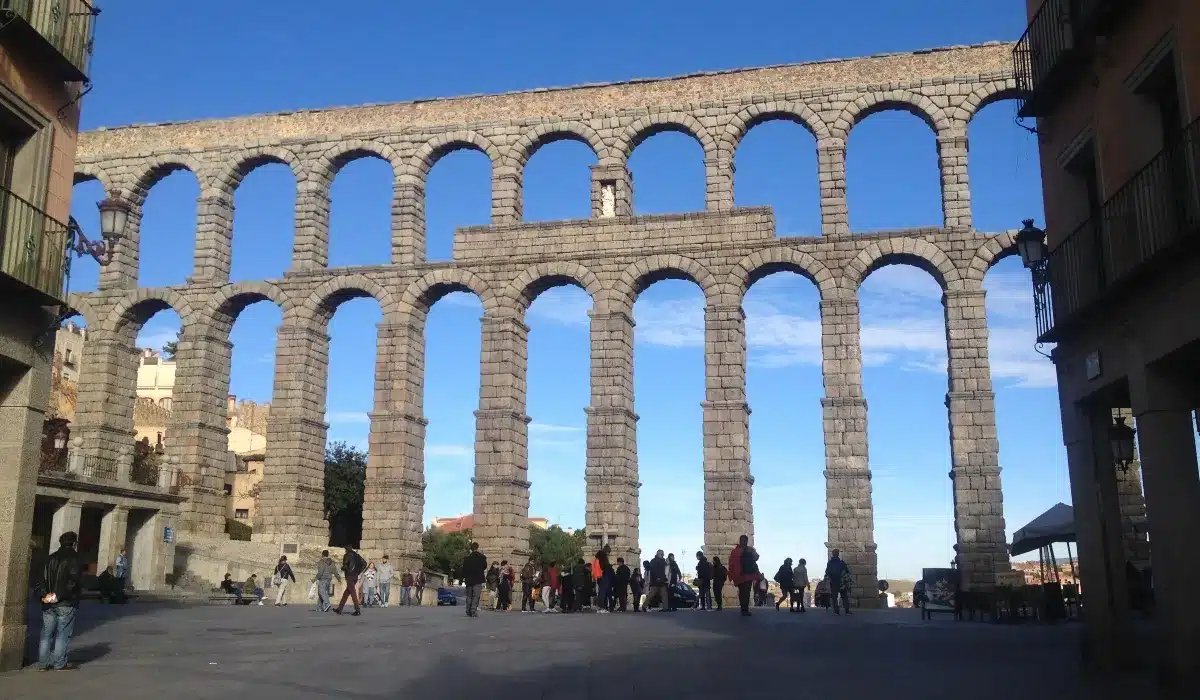 Vista frontal del Acueducto de Segovia en la plaza del Azoguejo, con turistas paseando bajo sus arcos romanos.