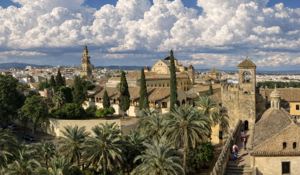 Vista panorámica de Córdoba con la Mezquita-Catedral y el Alcázar, palmeras y cipreses en primer plano bajo un cielo azul con nubes naturales y luz profesional