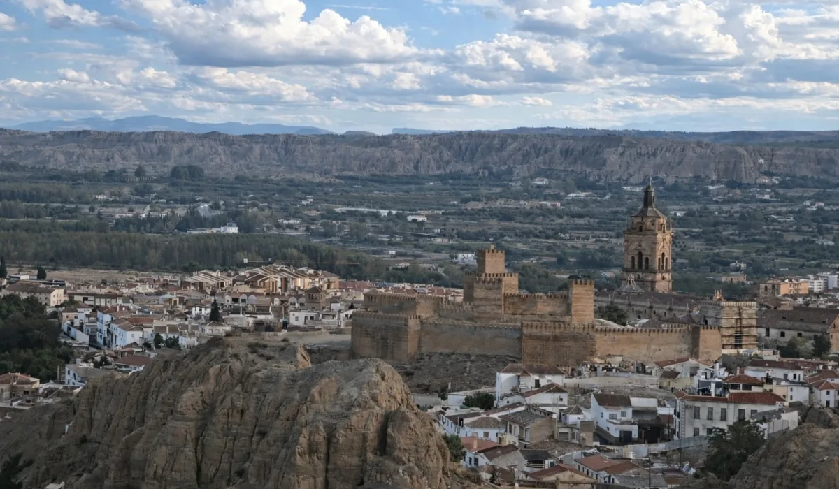 Vista panorámica de Guadix con la Alcazaba y la torre de la catedral al atardecer, casas blancas entre formaciones rocosas y cielo azul con nubes.